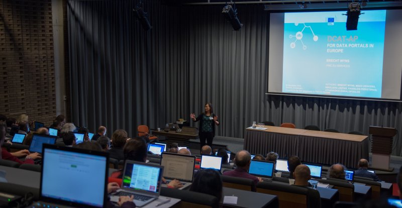 Facing the front of the lecture theatre. A woman addresses the audience, the large screen is ready for the DCAT-AP presentation.