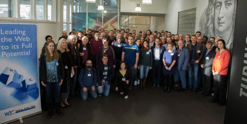 A group shot of roughly 80 people in a hall. A portrait of Sir Isaac Newton can be seen on the wall on the right