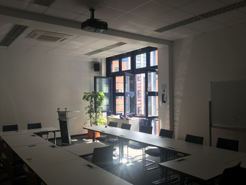 Inside of a meeting room with conference tables arranged roughly in a U-shape and about 10 chairs visible in the photo, and in the far corner there are open double-door large windows letting in natural light and outside air